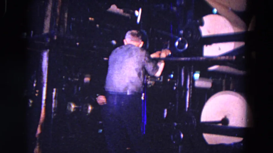 In 1967 Trenton, New Jersey metadata context, a worker appears beside heavy machinery with pale round drum-like parts in a dark factory space.