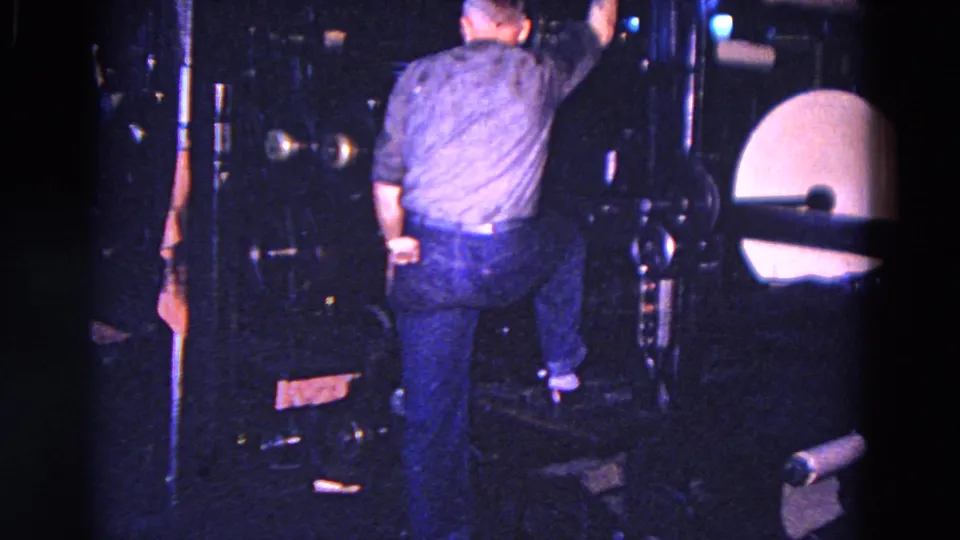 The worker is seen from behind while climbing or positioning himself on factory equipment, with large pale circular machine parts nearby.