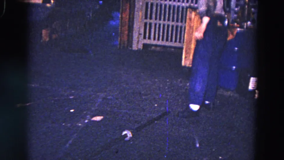 The worker stands beside industrial equipment in a dark interior while a loose cylindrical component remains visible on the floor.