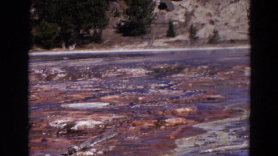 1967 Yellowstone 8mm still with reddish-brown geothermal deposits, pale mineral patches, water, trees, and hillside in the background.