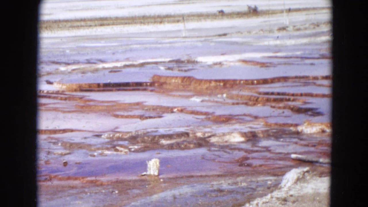 1967 Yellowstone 8mm frame focused on pale mineral flats, reddish terrace bands, shallow water, and geothermal crust.
