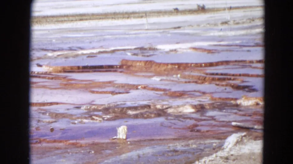 1967 Yellowstone 8mm frame focused on pale mineral flats, reddish terrace bands, shallow water, and geothermal crust.
