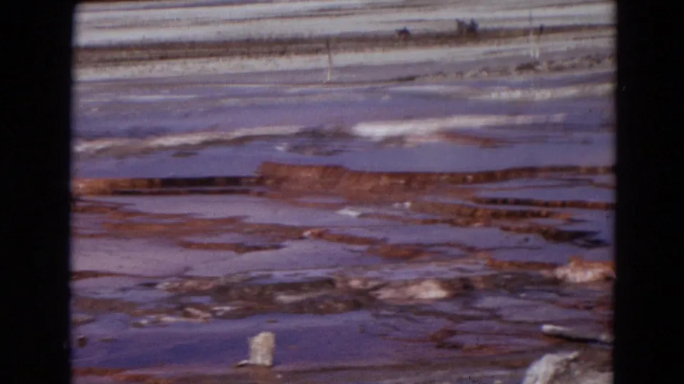 1967 Yellowstone 8mm frame showing shallow geothermal pools, reddish mineral terraces, white crust, and distant basin edge.