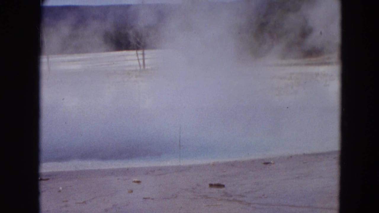 Steam drifting over a pale geyser basin with trees in the distance and dark film borders.