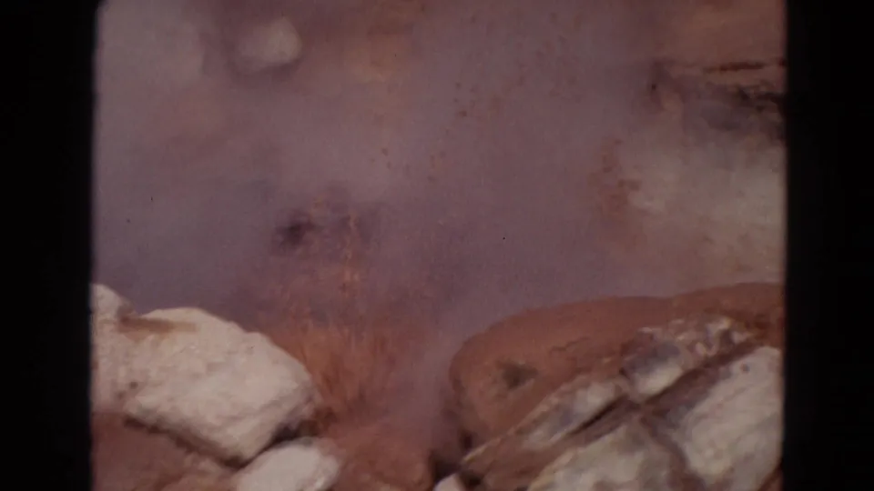 Close 1967 Yellowstone frame showing reddish mineral ground, pale rocks and a hazy geothermal vent area in small-gauge film texture.