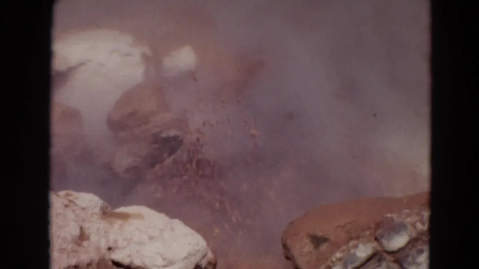 1967 Yellowstone 8mm frame showing a muddy geothermal burst behind foreground rocks, partly obscured by steam and film grain.