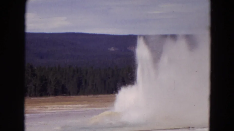 1967 Yellowstone 8mm frame showing a geyser erupting with white steam and water against forested hills and pale sky.