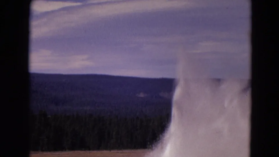 The 8mm frame holds on the Old Faithful eruption from a visitor viewpoint, with steam, forest, and distant hills visible.