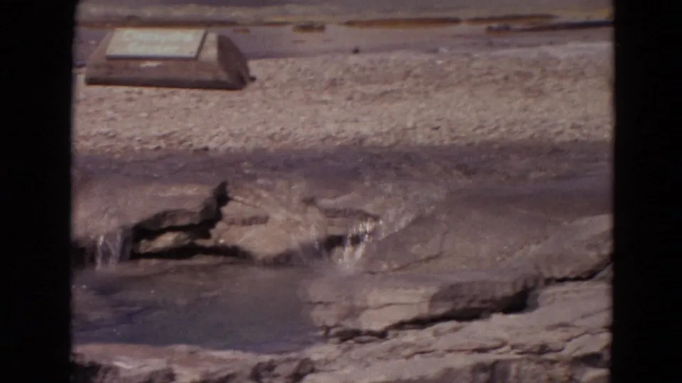 A nearby Yellowstone thermal feature appears with shallow water, mineral-stained ground, rocks, and a small sign or marker in the background.