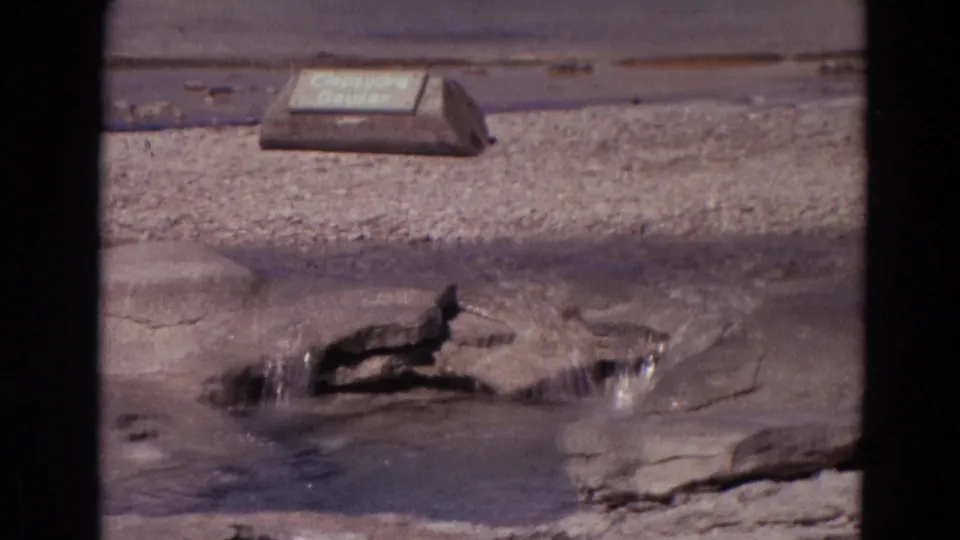 Clepsydra Geyser in Yellowstone appears in a 1967 8mm frame, with water gushing from rock and the identification sign nearby.