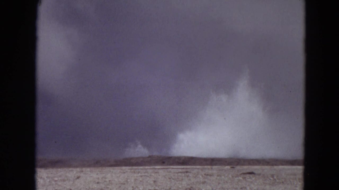 Distant white plume rising from a flat landscape beneath a dark overcast sky.
