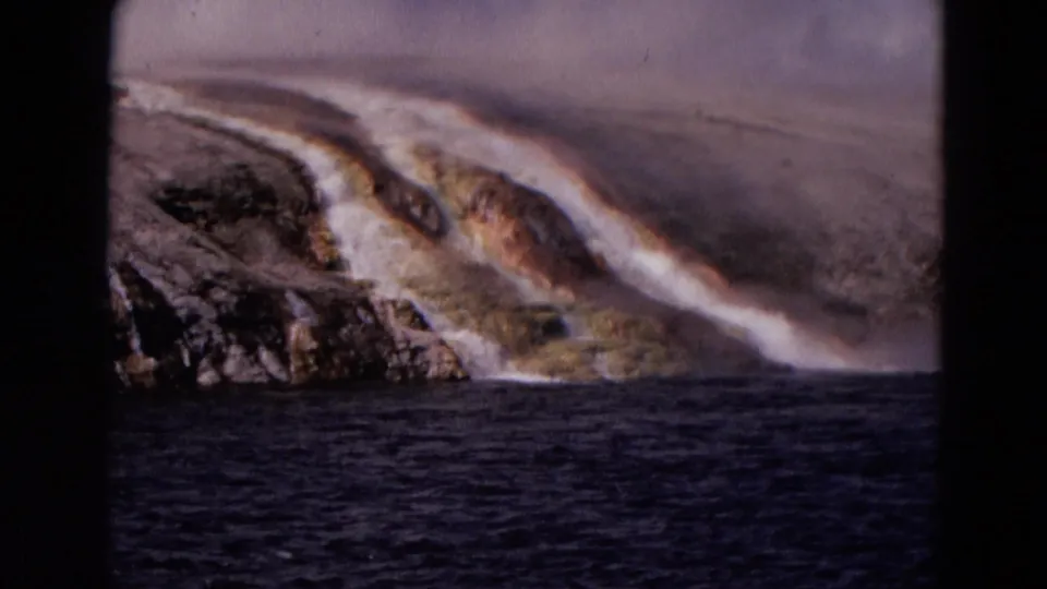 1967 Yellowstone 8mm frame of steaming mineral-streaked shoreline rock beside dark water.