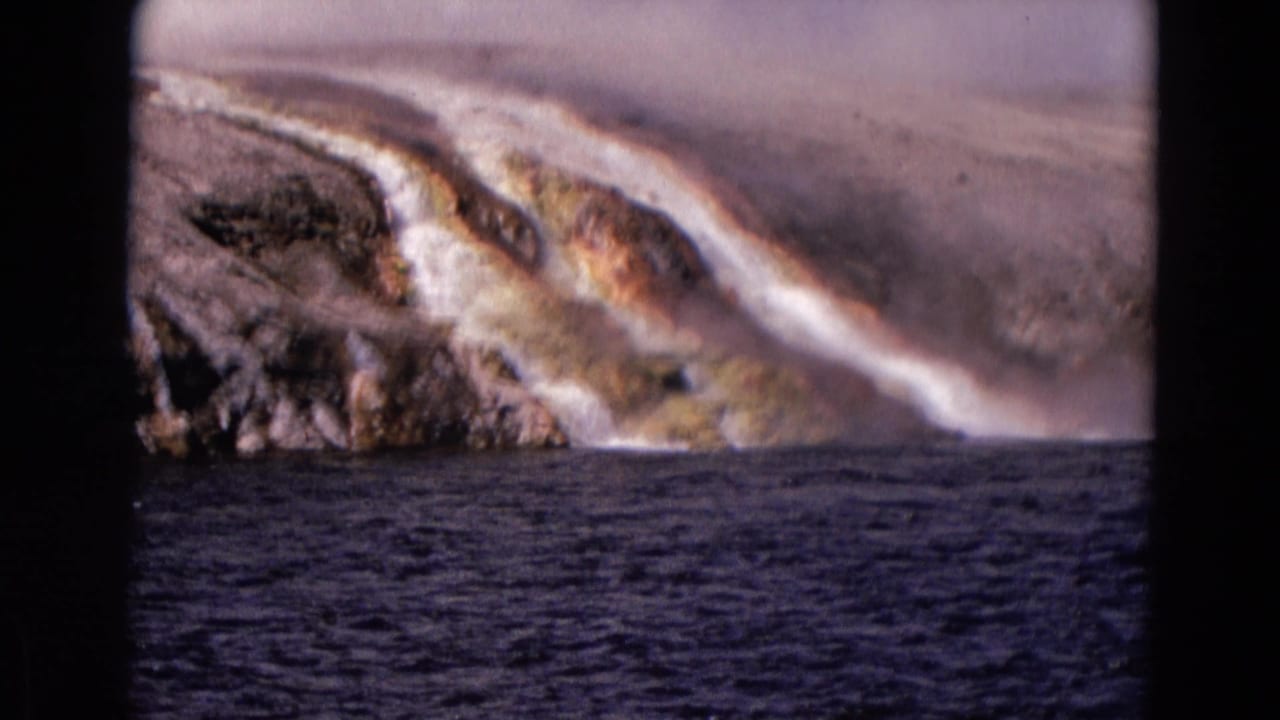 Steam rises over mineral-streaked runoff flowing down a rocky slope beside dark water.