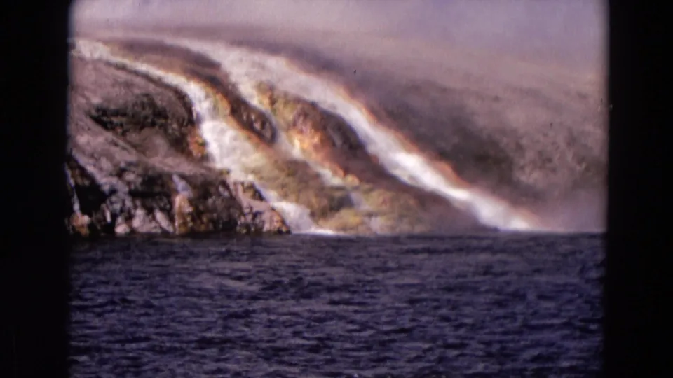 The frame holds on a Yellowstone geothermal slope with steam, white runoff, orange mineral deposits, and rippled dark water below.