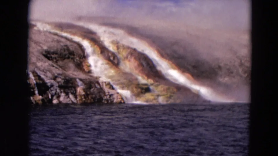 1967 Yellowstone home-movie footage shows pale mineral water trickling down rock with steam haze and dark water in the foreground.