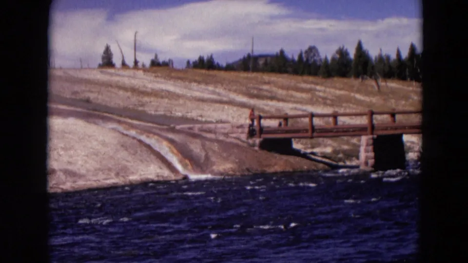 1967 Yellowstone frame showing a wooden footbridge over rushing water, pale banks, scattered trees, and a partly cloudy sky.