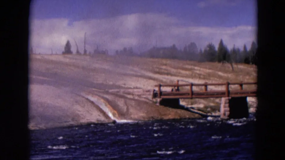 1967 Yellowstone home-movie frame with a wooden bridge, turbulent water, misty foreground, pale ground, and distant tree line.