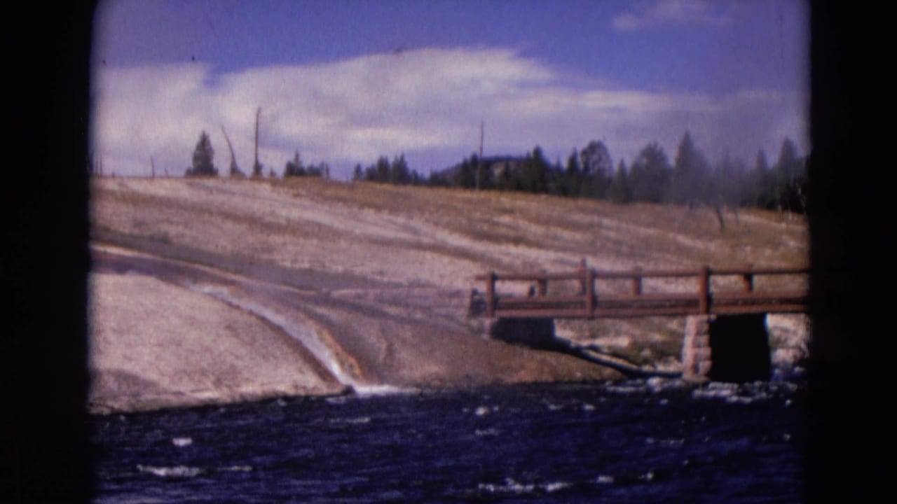 Wooden bridge over a dark river beside a pale sloped bank under blue sky.