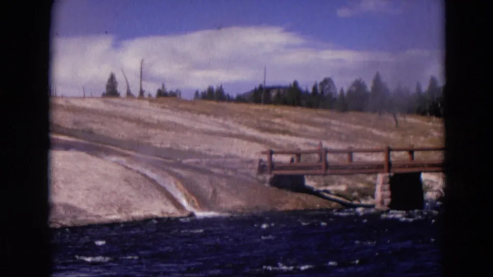 1967 Yellowstone home-movie frame showing a wooden bridge, dark water, pale mineral bank, trees, and clouds.