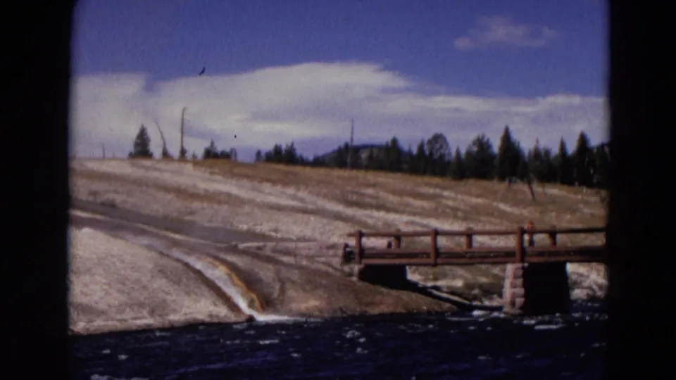 1967 Yellowstone home-movie view of a bridge and river with pale ground, trees, and wind-bright clouds.