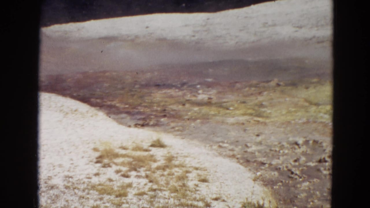 1967 Yellowstone 8mm frame with a pale mineral channel curving past a hazy geothermal basin and distant terrain.