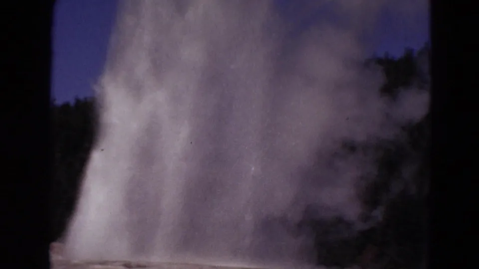 1967 Yellowstone 8mm frame showing a geyser eruption sending water and steam upward beside dark trees under blue sky.