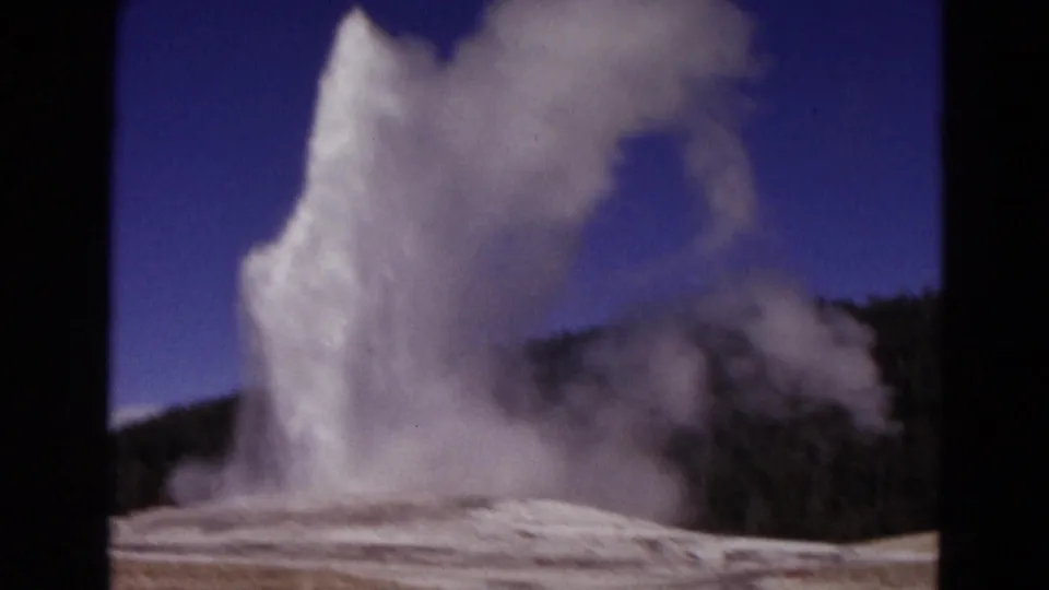 Visitor-shot 1967 Yellowstone frame with a tall geyser column, drifting steam, pale ground, forest ridge, and deep blue sky.