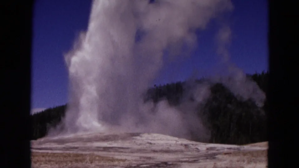 1967 Yellowstone 8mm still showing a geyser plume leaning upward, with steam drifting over the basin and forested hills behind.