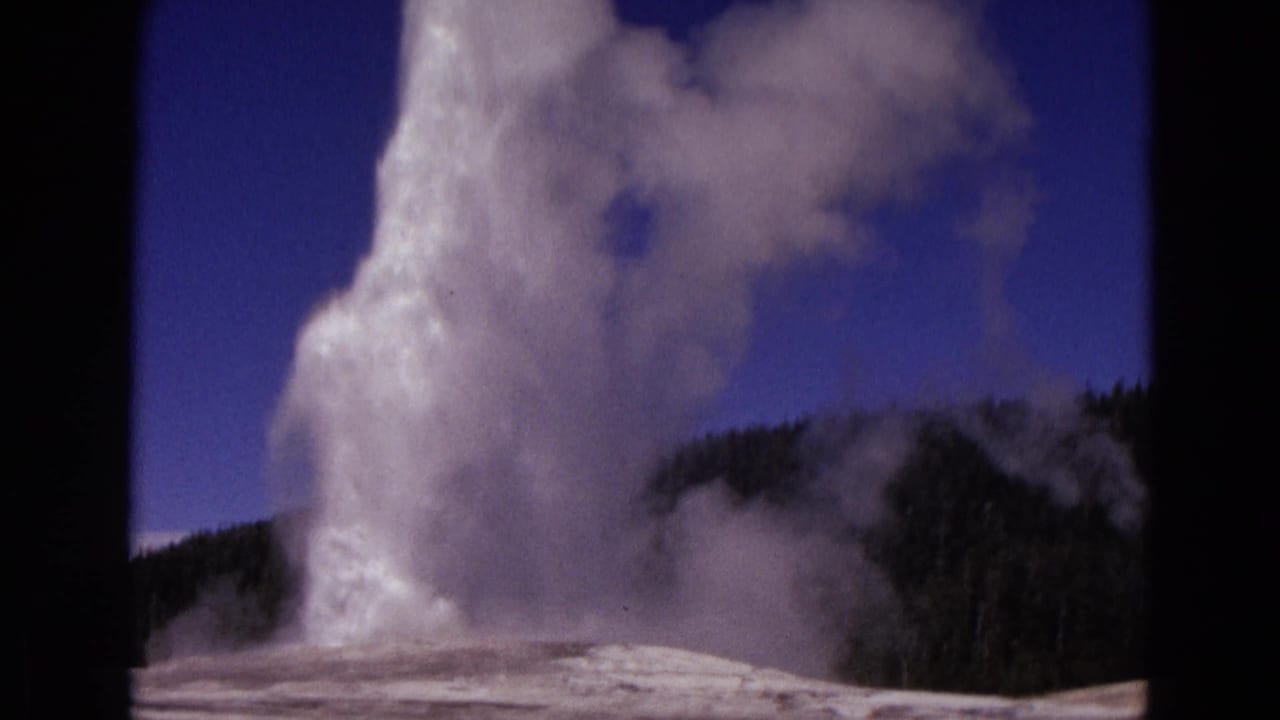 Old Faithful geyser erupts against a blue sky with pine-covered ridge behind it.