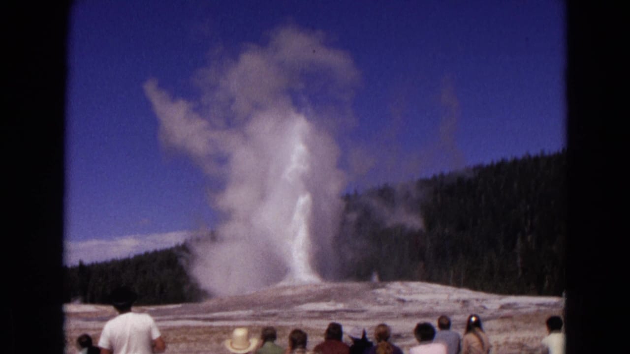 Old Faithful erupts behind tourists standing and seated near the geyser basin.