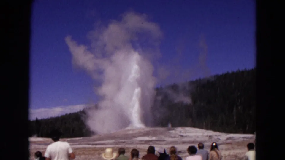 1967 8mm Yellowstone footage shows Old Faithful erupting as tourists watch from the geyser basin under a blue sky.