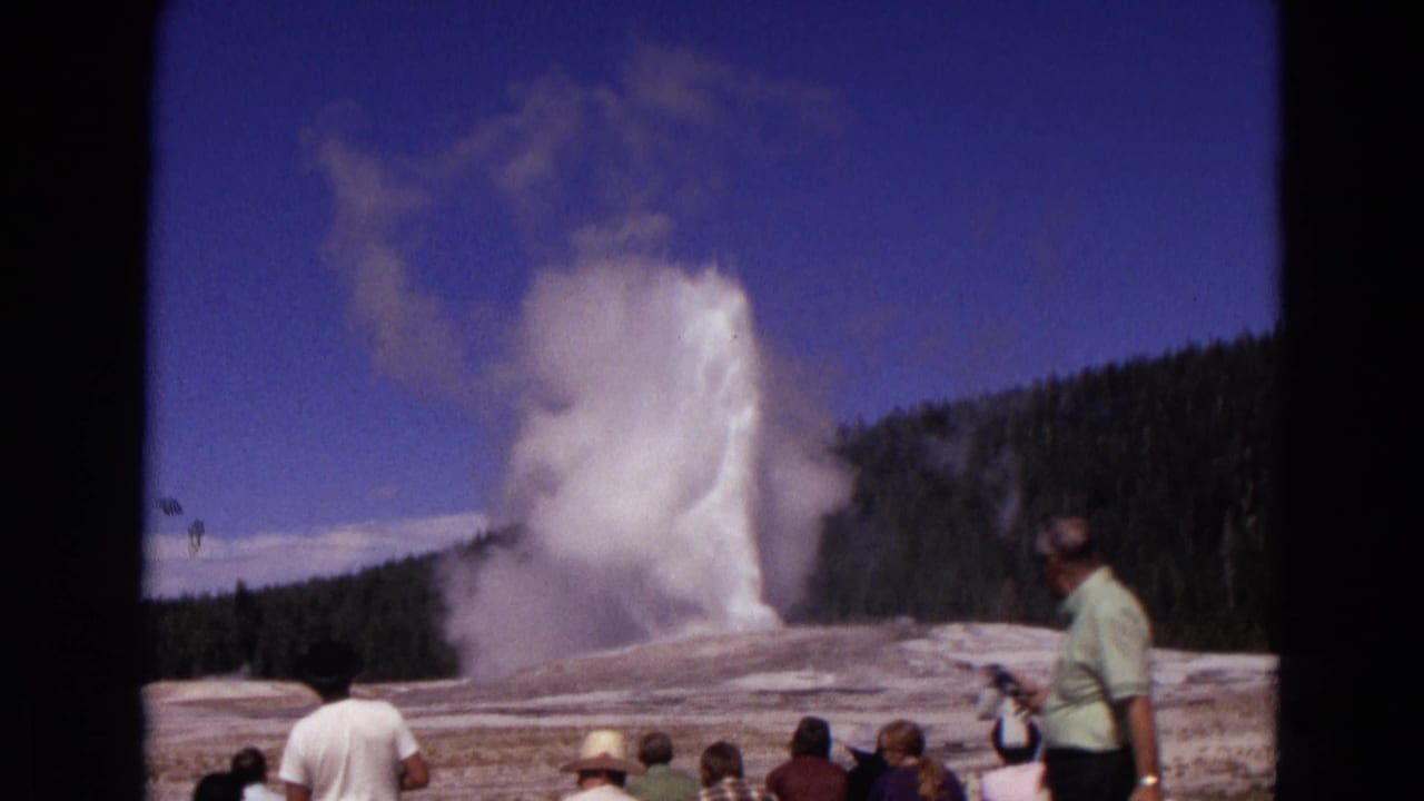 A tall geyser plume rises while tourists watch, including a man standing at right.