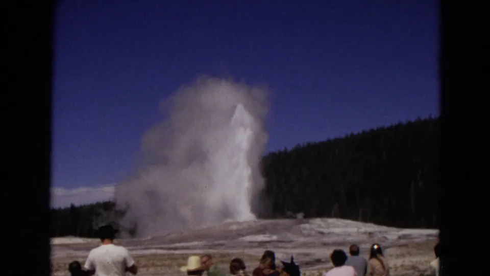 The Old Faithful eruption continues, with a thick steam cloud drifting across the basin while tourists remain in the foreground.