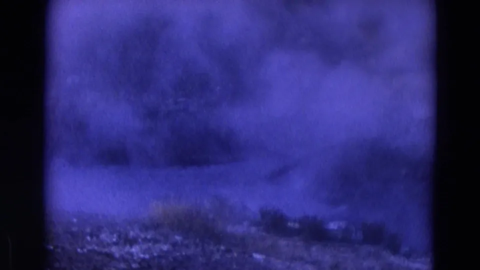 1967 Yellowstone home-movie frame with heavy blue haze or smoke, faint trees, and uneven rocky ground.
