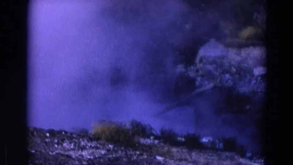 1967 Yellowstone home-movie frame where smoke or steam fills the foreground above gravel, rocks, and small grass clumps.
