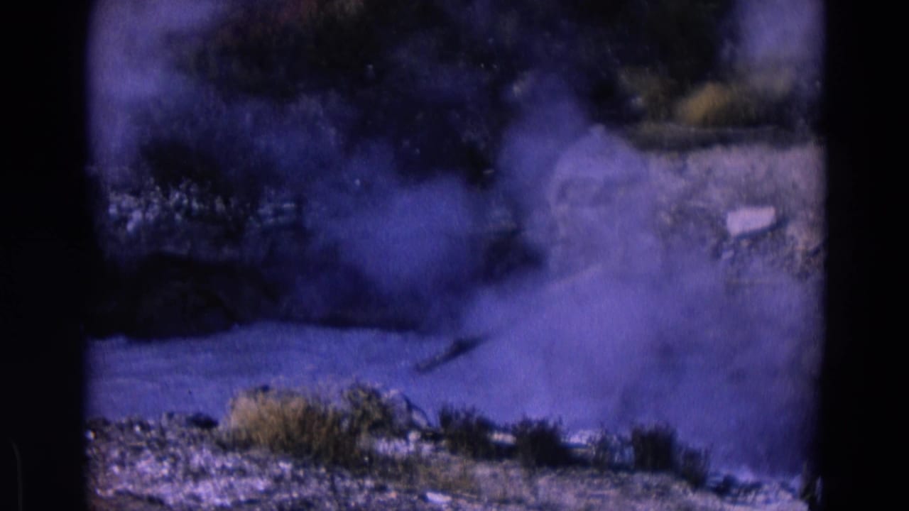 Steam drifts across a rocky vent area with brush and a pale pipe-like line.