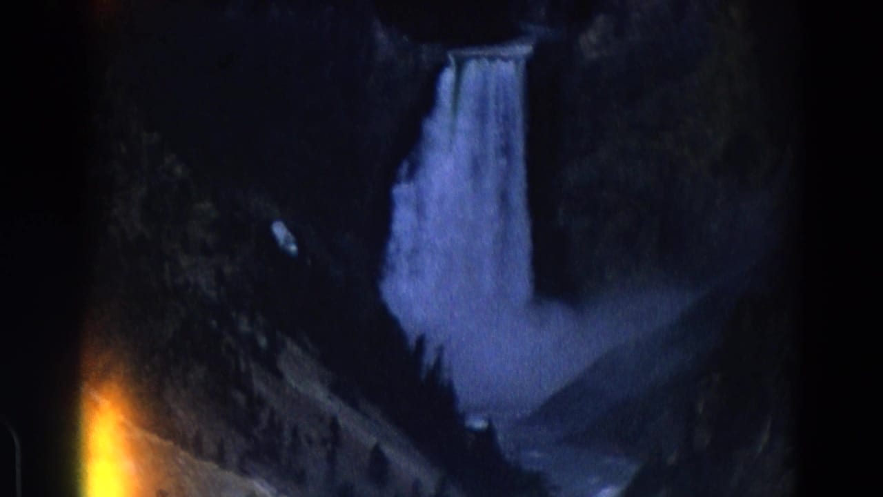 Distant waterfall dropping through a dark rocky canyon with film edge flare at lower left.