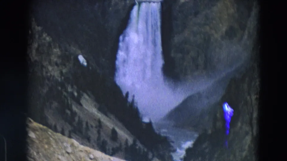 1967 Yellowstone 8mm frame showing the waterfall, canyon slopes, rocky foreground, film border, and a blue scan or film artifact at right.