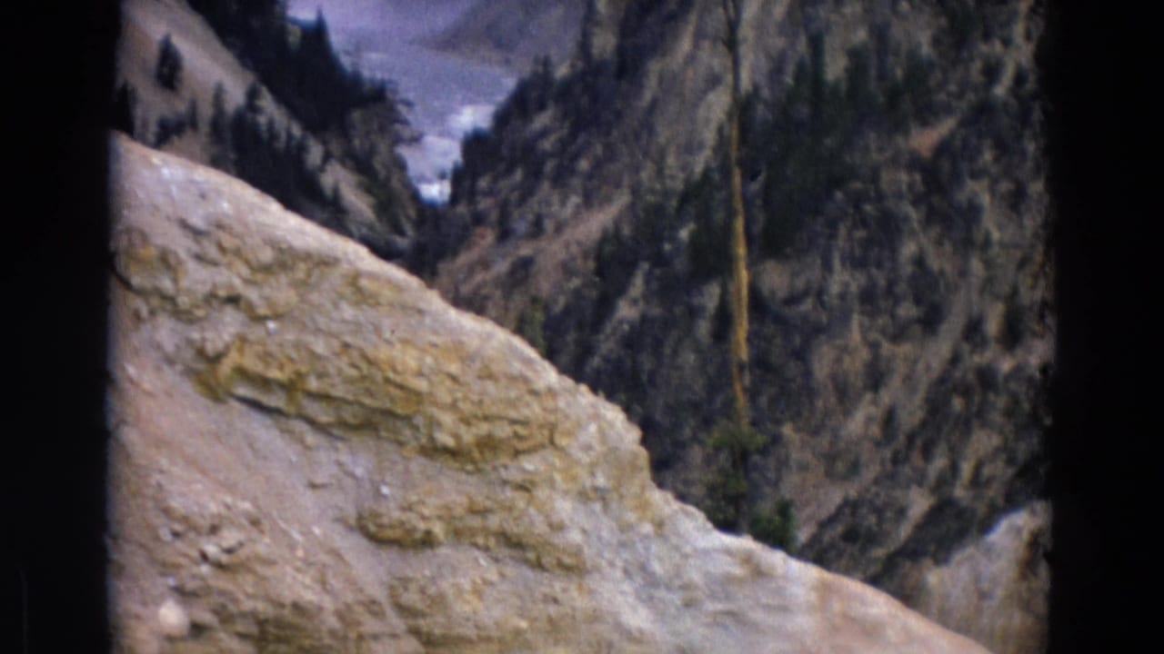 Rocky canyon ledge with steep cliffs, trees, and distant water between the slopes.