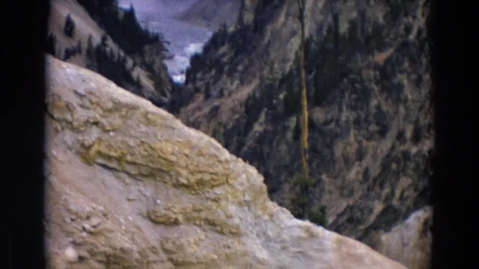 1967 Yellowstone frame with a pale foreground ledge, rugged canyon walls, scattered evergreens, and a glimpse of water in the distance.