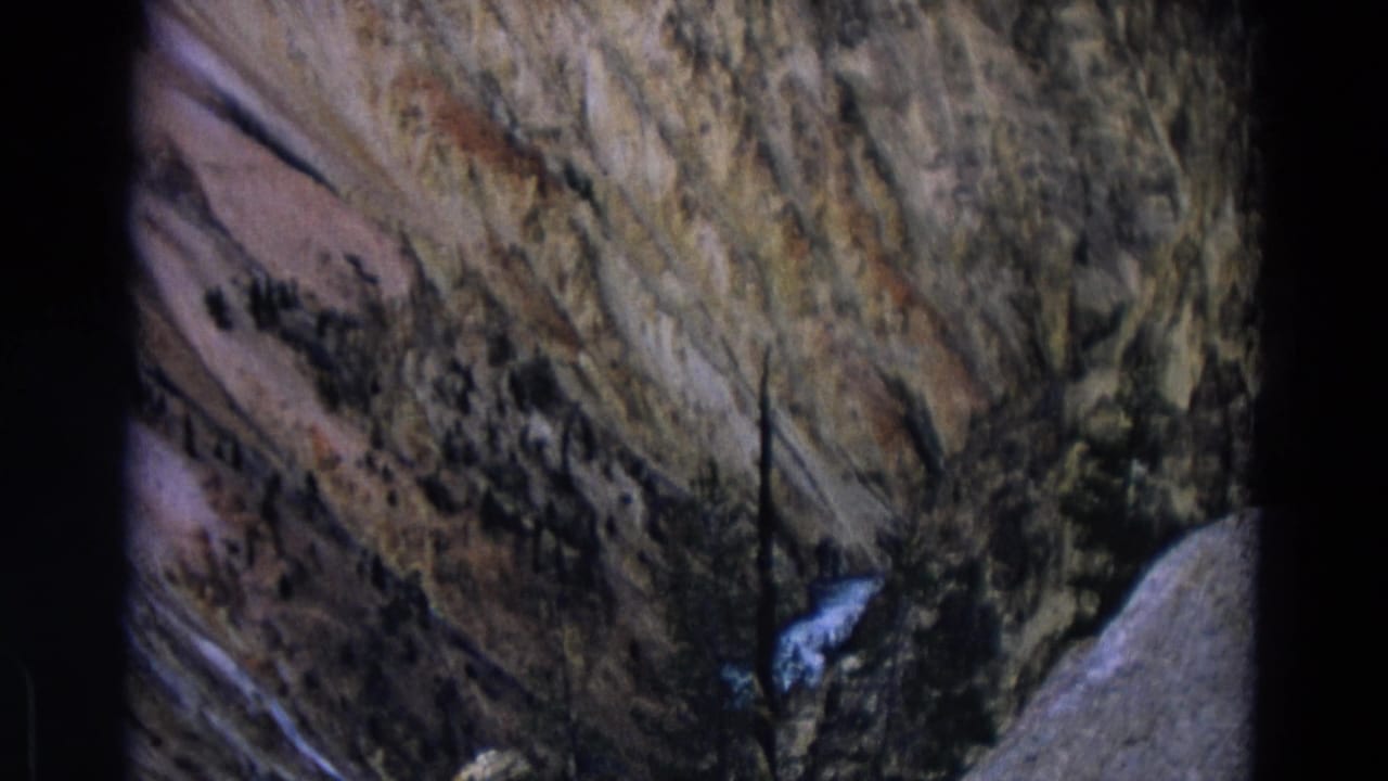 Steep tan rock slope with a small bright waterfall and sparse trees.