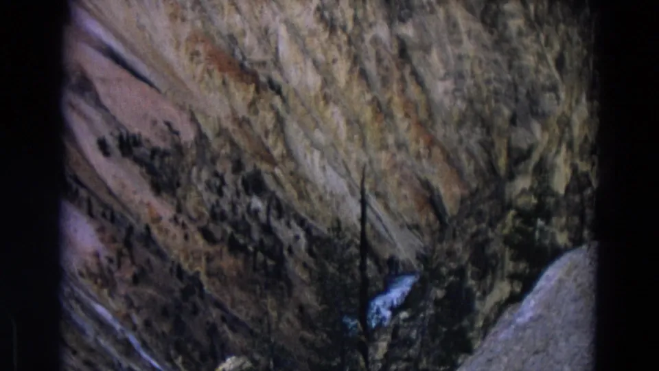 Yellowstone 1967 frame showing a small cascade against layered canyon rock, with scattered evergreen trees and faded 8mm color.