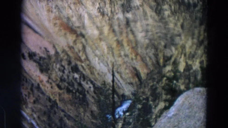 1967 Yellowstone 8mm frame of a rugged canyon wall, sparse trees, and a small section of white water near the bottom of the view.
