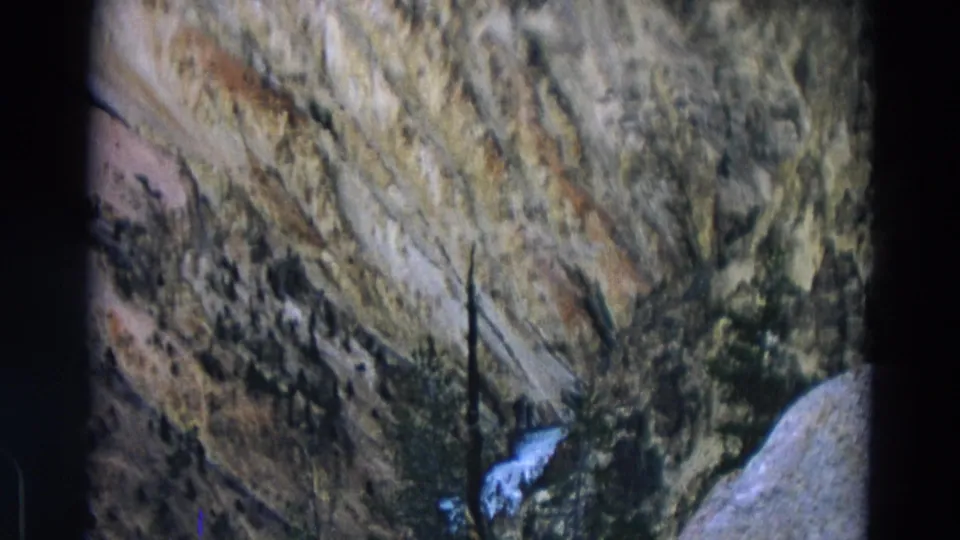 Faded 8mm Yellowstone frame showing a rocky canyon slope, scattered evergreens, and a small waterfall below the central rock face.