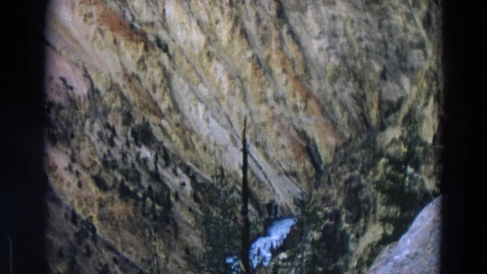 1967 Yellowstone home-movie frame showing a striated canyon wall, sparse evergreens, and a narrow white waterfall or stream.