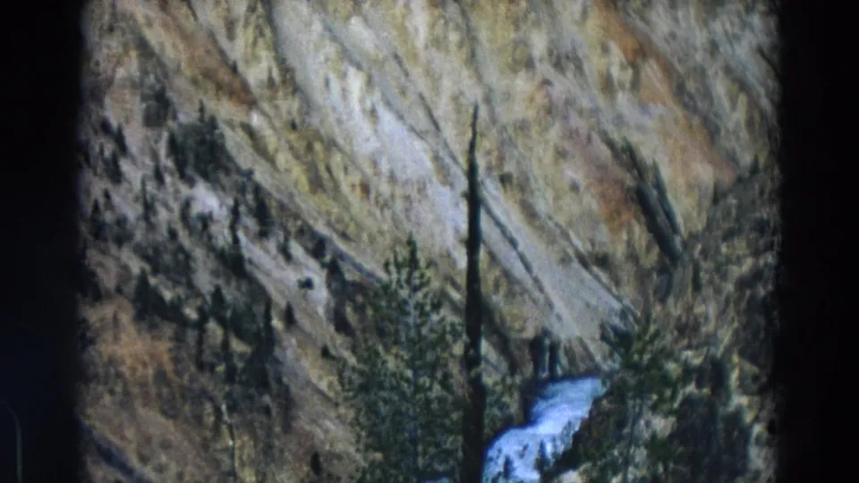 Vintage 8mm Yellowstone view of a steep rocky hillside, scattered evergreens, and a small stream or waterfall near the bottom.