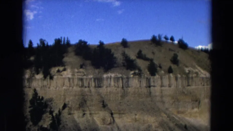 1967 Yellowstone home-movie frame showing a rugged layered cliff face, scattered evergreens, and a bright blue sky.