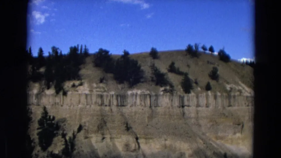 Vintage 8mm Yellowstone view of a sunlit cliff face, dry slope, and sparse evergreens beneath blue sky.
