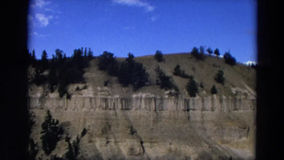 1967 Yellowstone home-movie frame showing a layered rock wall, tree-lined ridge, and saturated blue sky.