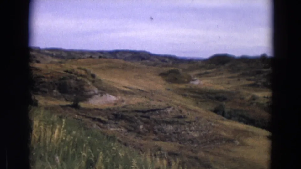 1967 Yellowstone home-movie frame showing dry rolling hills, exposed rock, scattered shrubs, and a distant low ridge.
